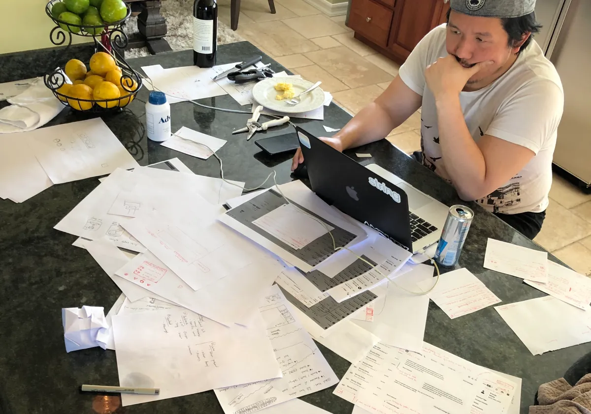 Design printouts and sketches spread across a kitchen table while a teammate reviews them on a laptop